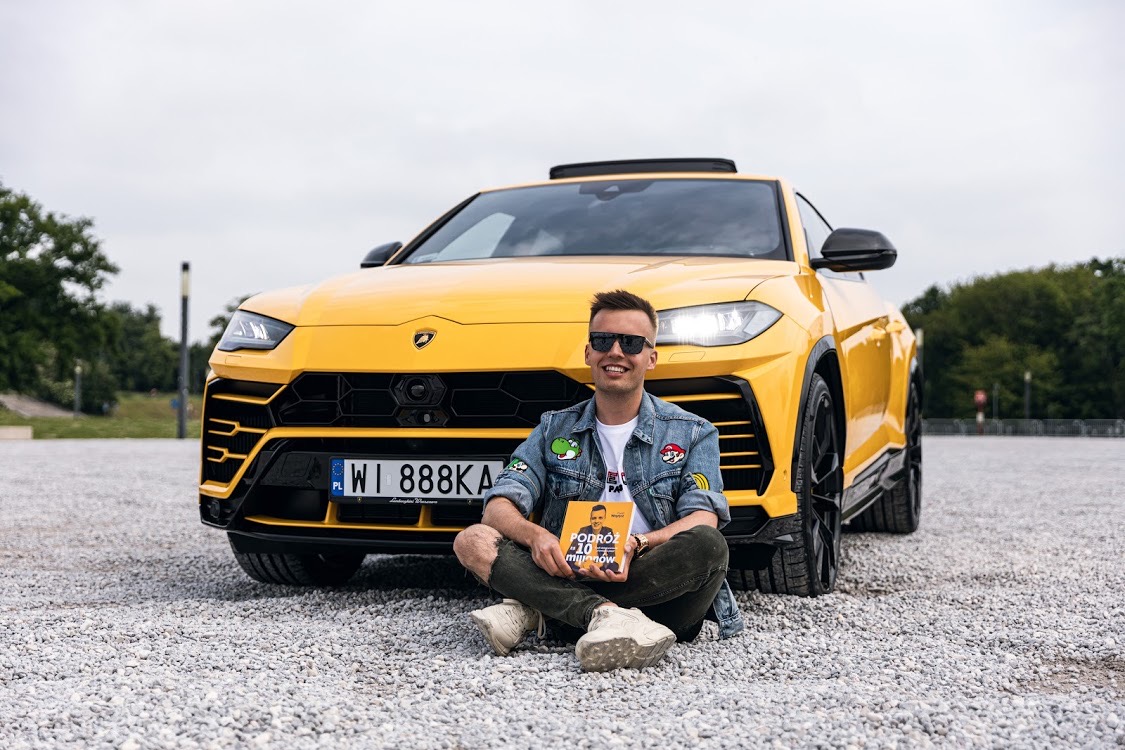 Paweł Wojnicz siedzący przy żółtym samochodzie trzymając swoją książkę